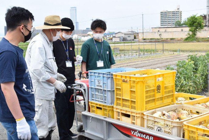 畑の前で作物を見つつ話を聞く男性